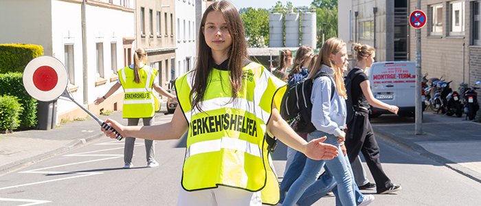 Schülerlotsin steht auf einer Straße, hinter ihr gehen einige Personen entlang.